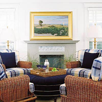 две windows flank each side of the fireplace with a gray hearth. Also, white walls brighten up the space with yellow and blue large plaid cushions on wicker arm chairs.