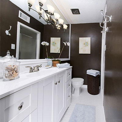 Дълбок chocolate walls of this renovated bathroom offset the crisp, white bathroom cabinets and the large, expansive bath mirror with a light fixture