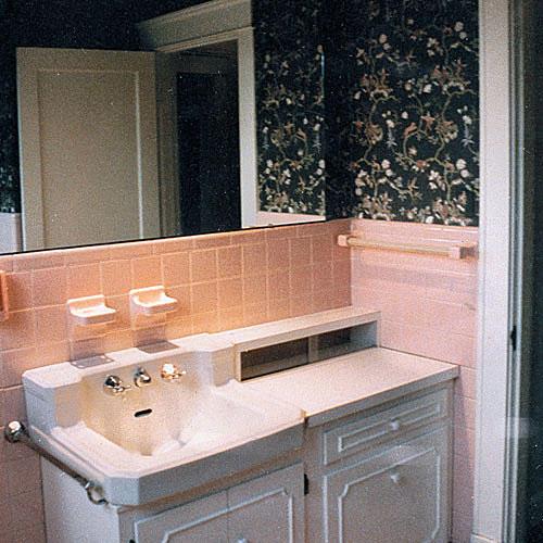 а before photo of a bathroom with pink tile around the sink area and dark, black flowered wallpaper