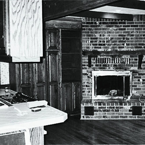 а before photo of a kitchen space with an island cabinet to the left, brown built-in shelving in the middle and an older fireplace off of the main section of the kitchen
