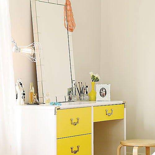 a rectangular glass-framed mirror sits atop a yellow desk with a variety of perfume, make up brushes, photo frames and jewelry decorate the top of it