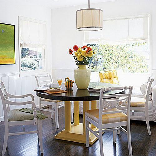 A bright, white dining room with crisp, white cloth shades over the windows and a large circular wooden dining room table with pale, yellow legs and white chairs surround it