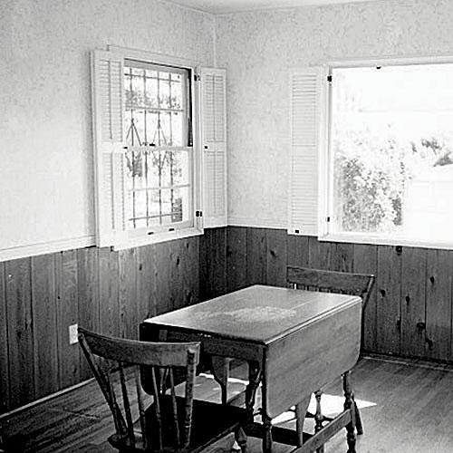 A before photo of a dining room with pine boards up half of the wall that has windows with interior shutters. A small table with two chairs is positioned in the middle of the room.